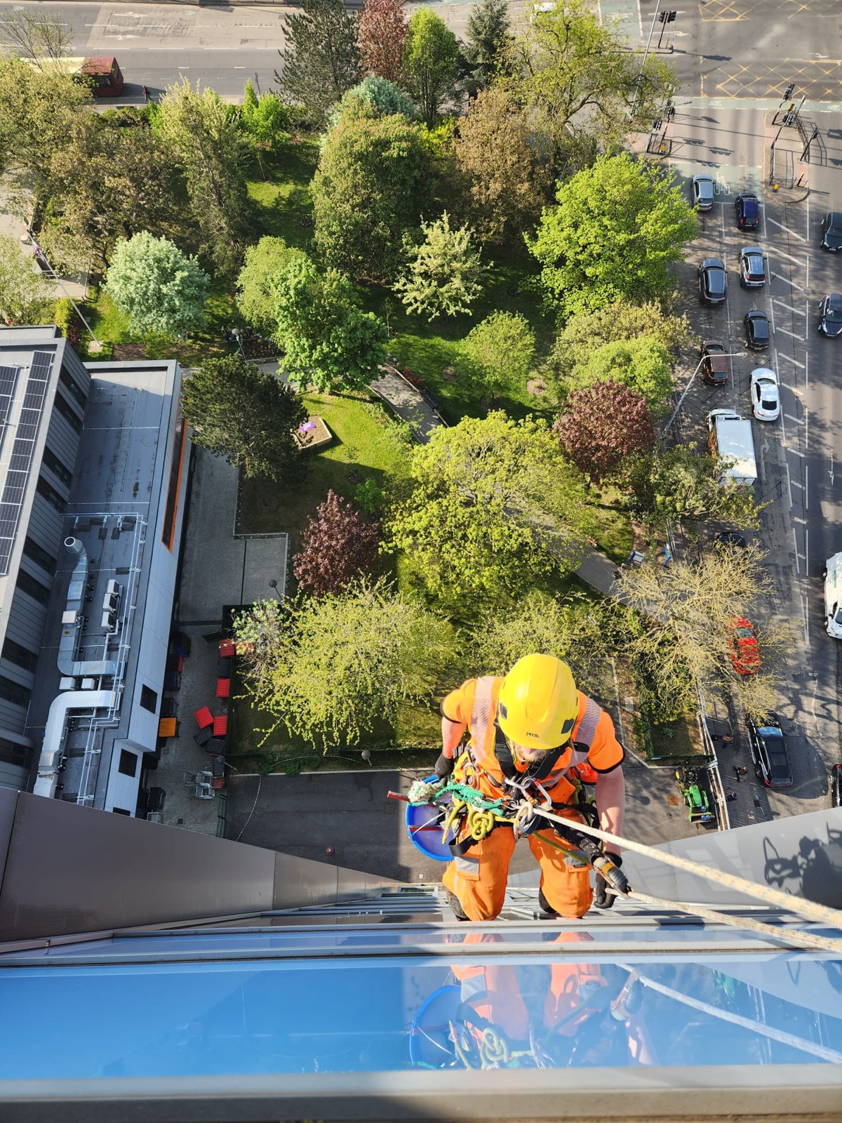 Ropesafe technician in orange PPE going over a roof edge, high above trees and a city road